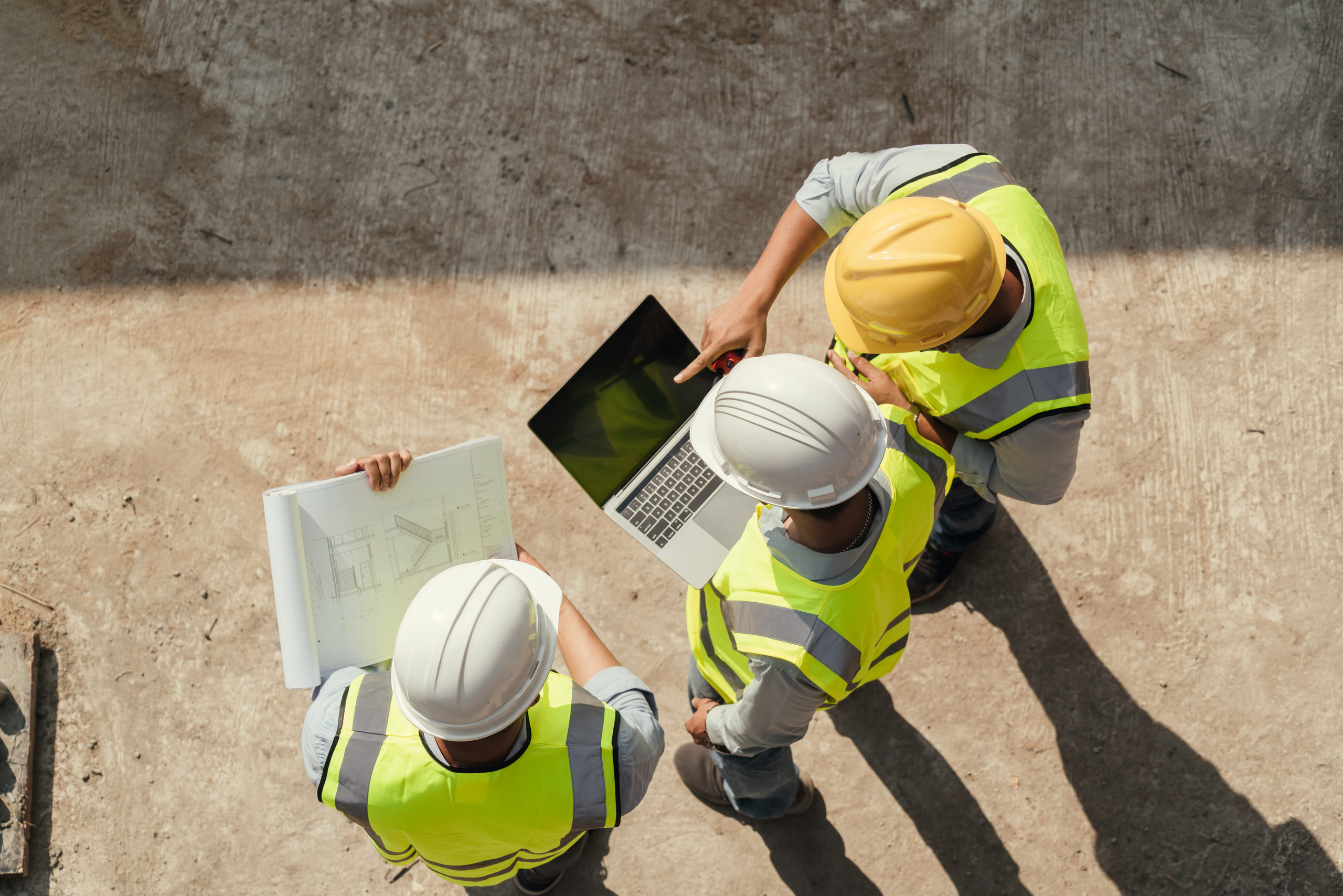 Vue de dessus de trois ingénieurs en gilet de sécurité fluo et casque de chantier consultant un ordinateur portable et des plans sur un chantier