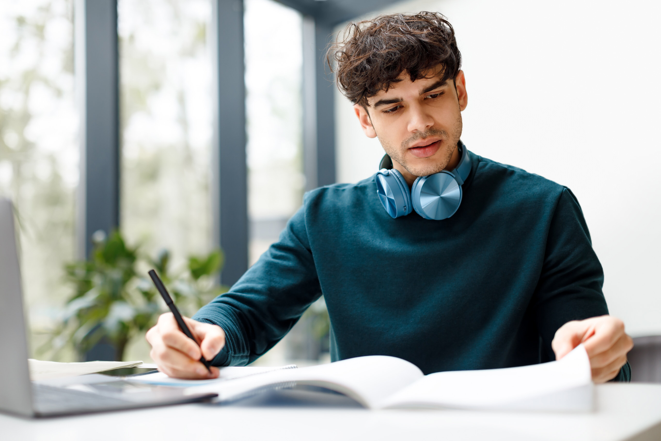 Étudiant concentré prenant des notes dans un cahier devant un ordinateur portable, avec un casque audio autour du cou