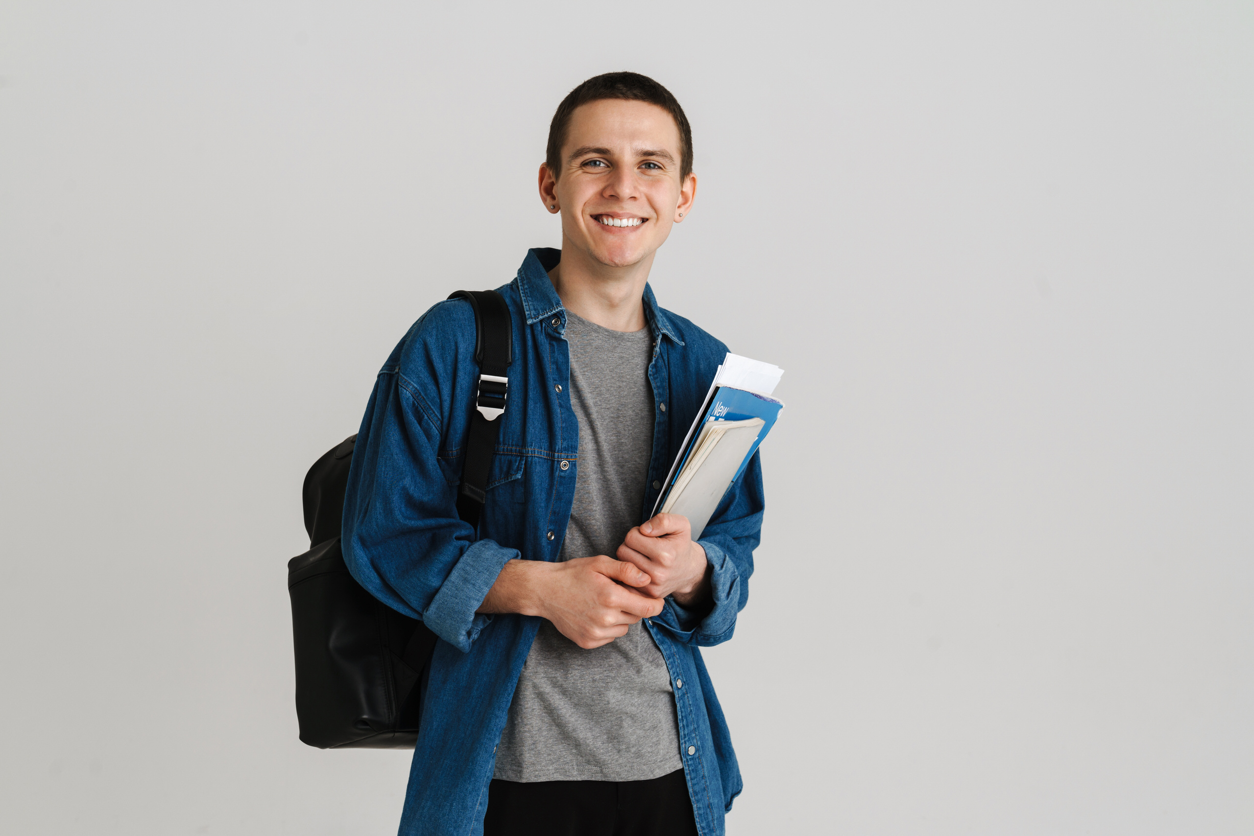 Étudiant souriant en chemise en jean portant un sac à dos et tenant des dossiers de cours