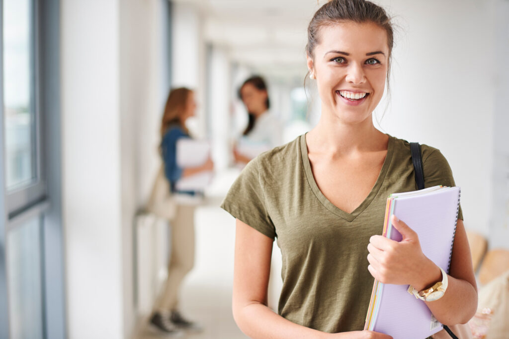 Étudiante souriante tenant un classeur dans le couloir d'un établissement, avec d'autres élèves en arrière-plan