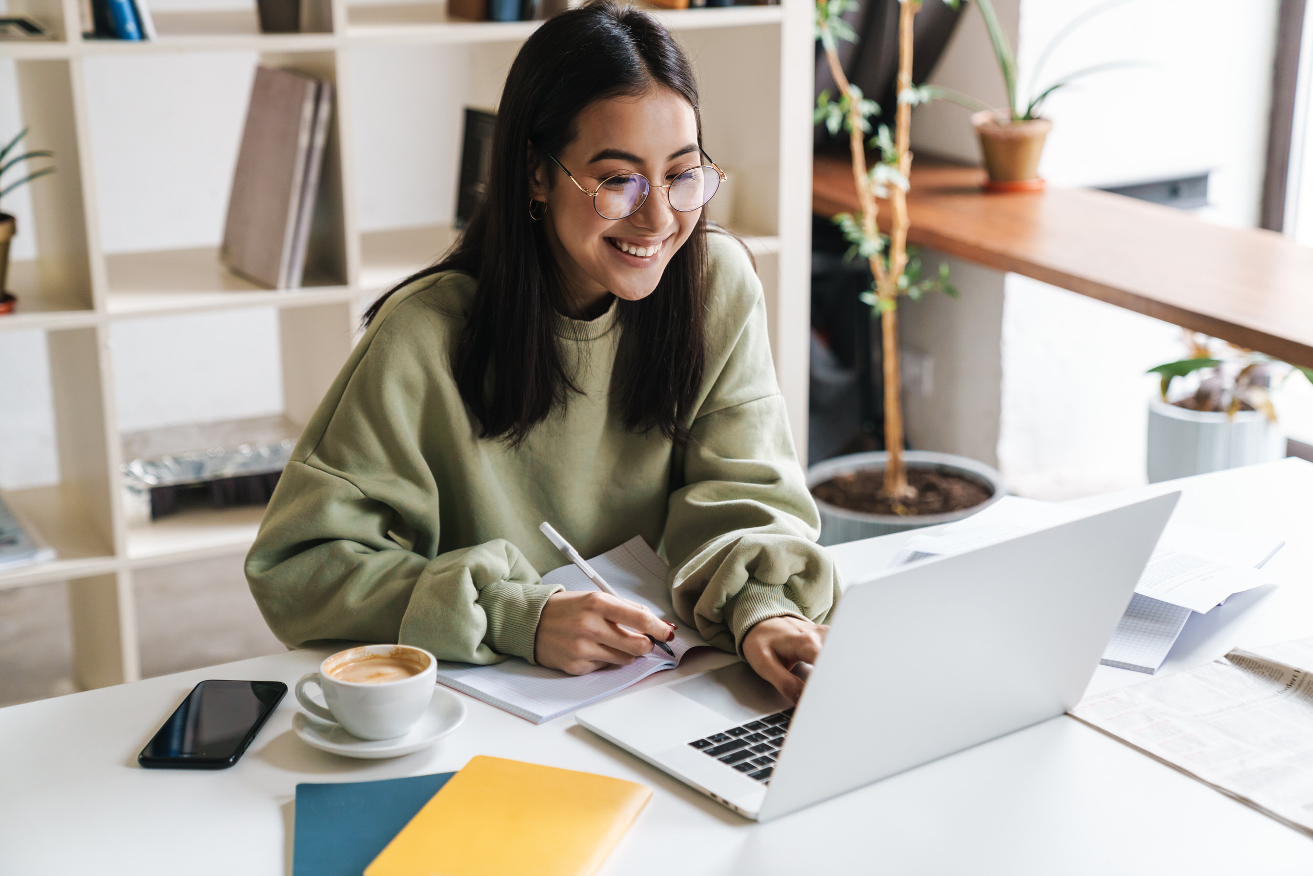 Étudiante souriante prenant des notes tout en travaillant sur son ordinateur portable, une tasse de café posée sur le bureau