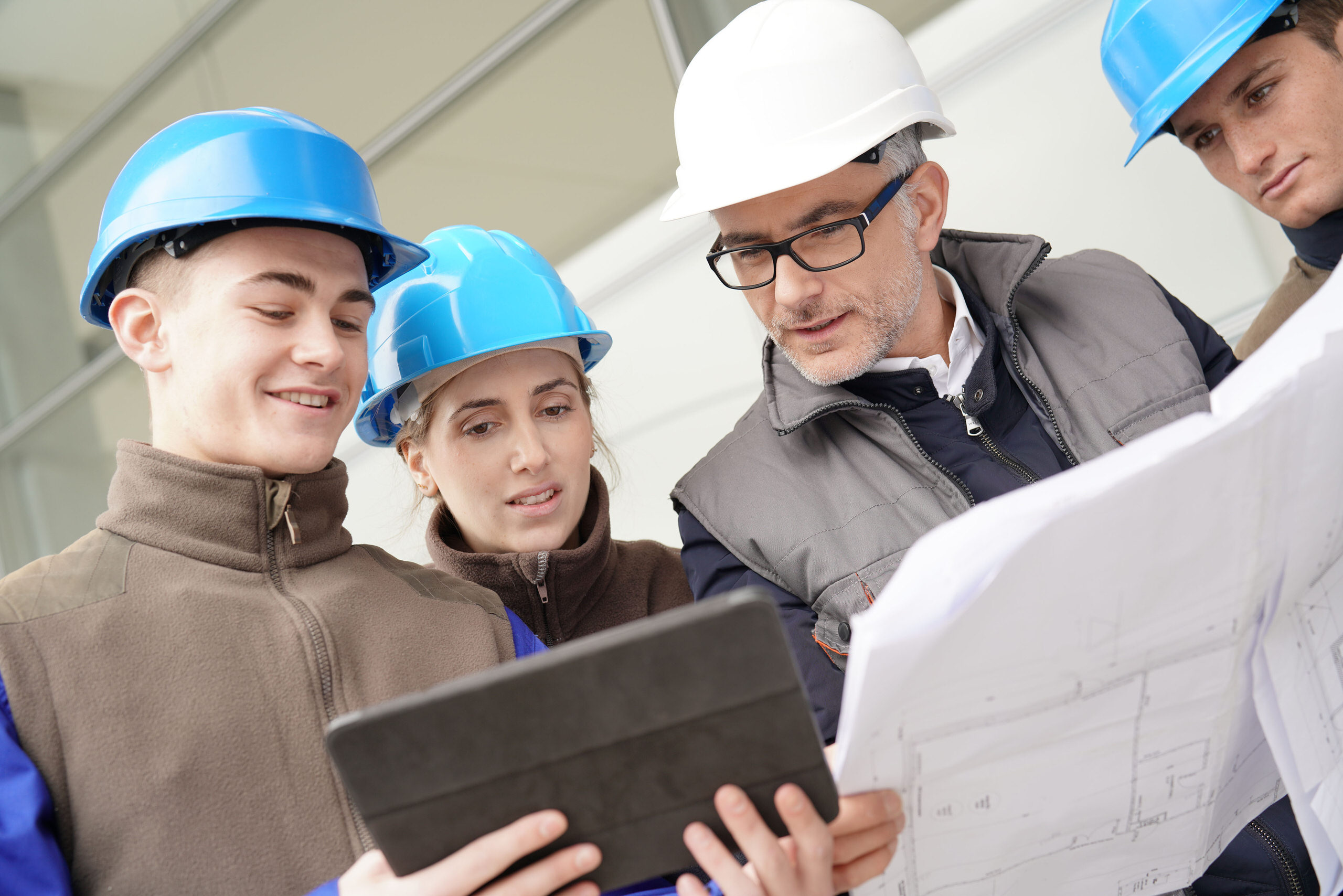 Formateur en casque blanc expliquant un plan à trois jeunes en formation portant des casques de chantier bleus, autour d'une tablette