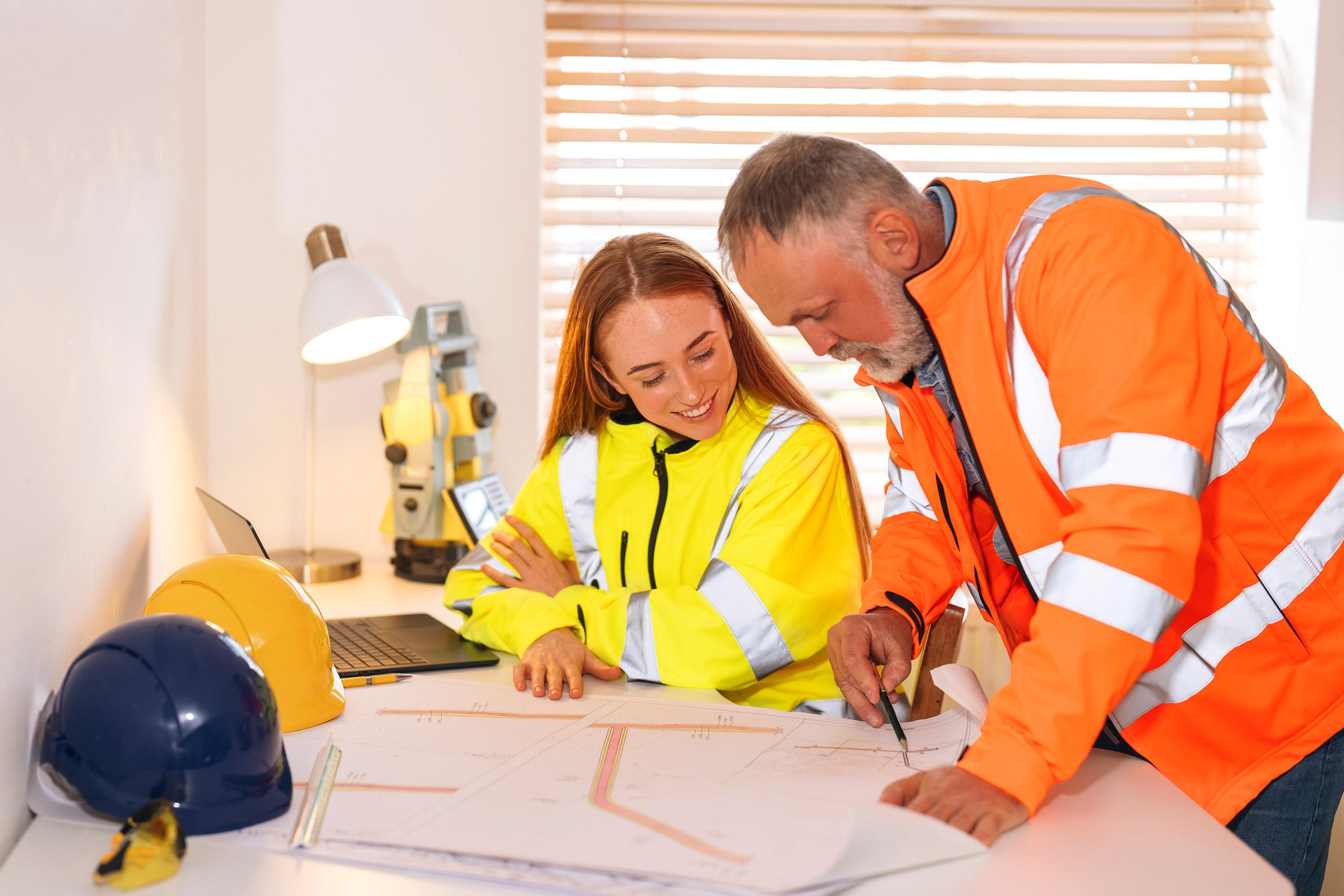 Étudiante en gilet de sécurité jaune et formateur en gilet orange examinant ensemble des plans de génie civil avec un appareil de topographie à proximité